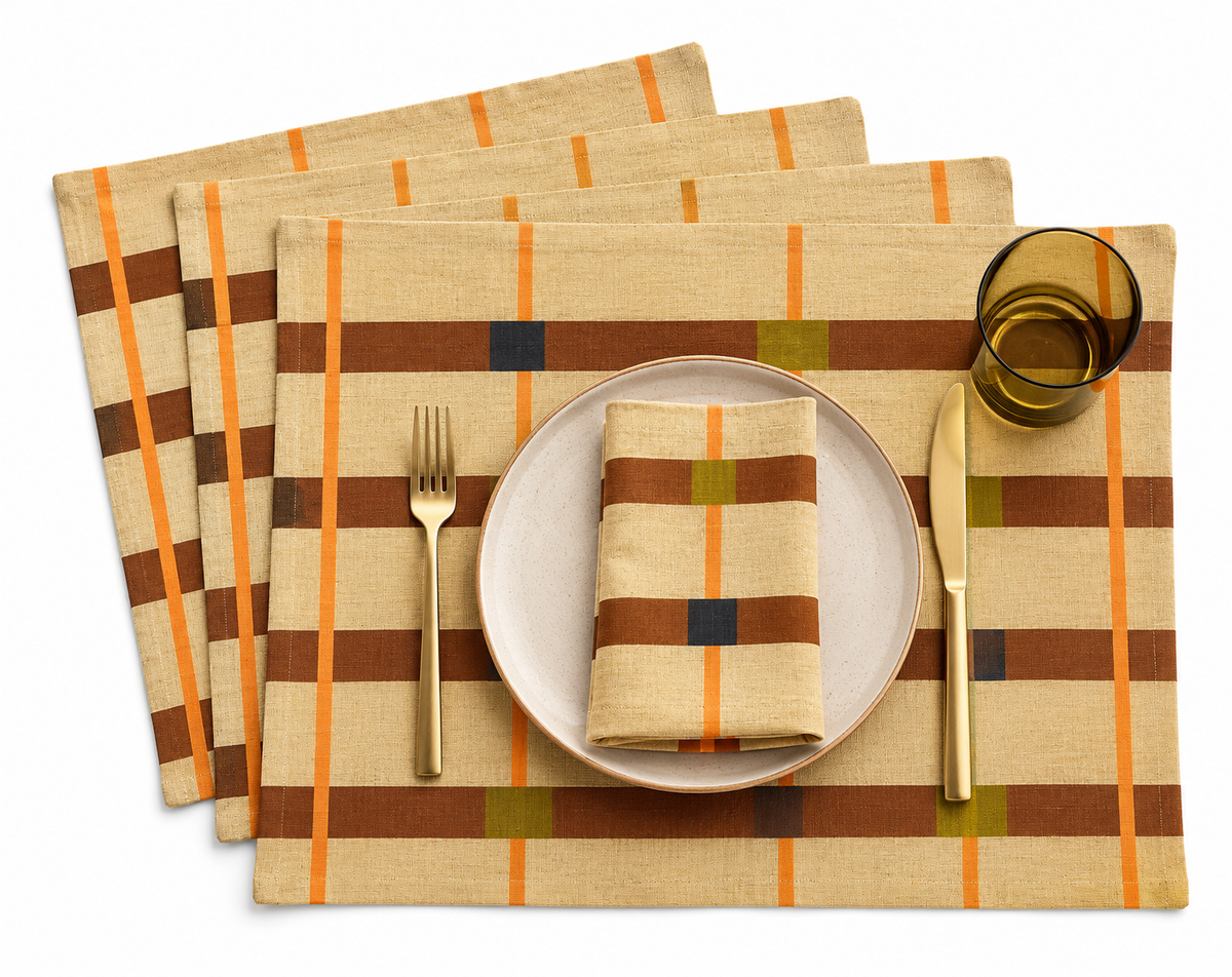 A Fields designer placemats by Lulasclan shows a set of four digitally printed linen and cotton blend placemats with a checkered pattern set against a white background. The design features rust brown horizontal stripes, bright orange vertical stripes, and small blue and ochre square accents. Styled with a gold fork and knife cutlery placed besides a white plate, the forth placemats is placed on top of the plate and a ochre transparent glass in view