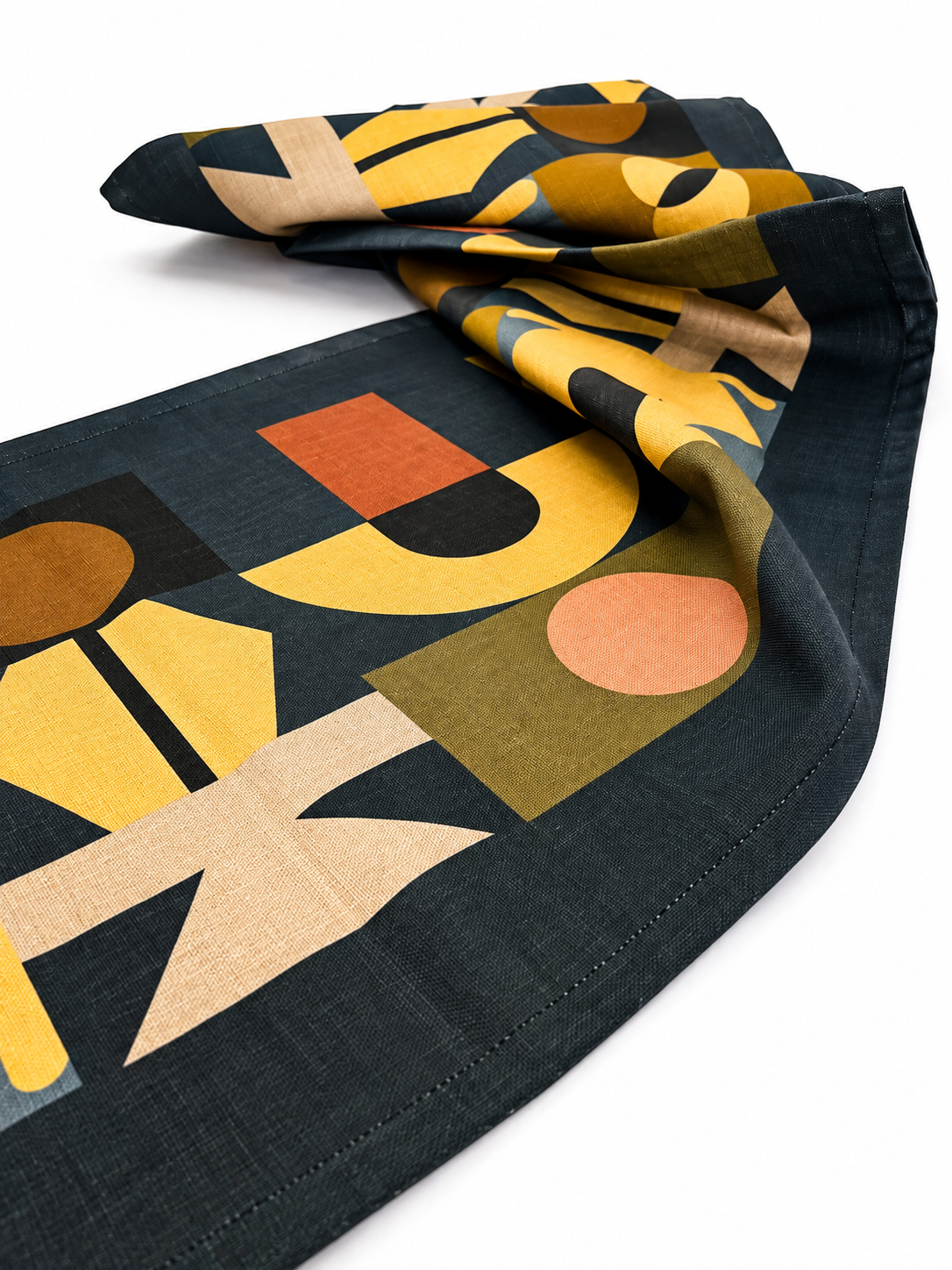 A Lulasclan The gathering table runner folded and draped scarf featuring a bold, abstract geometric pattern with mustard yellow, tan, terracotta, olive green, and peach shapes on a dark charcoal background, set against a clean white studio backdrop.