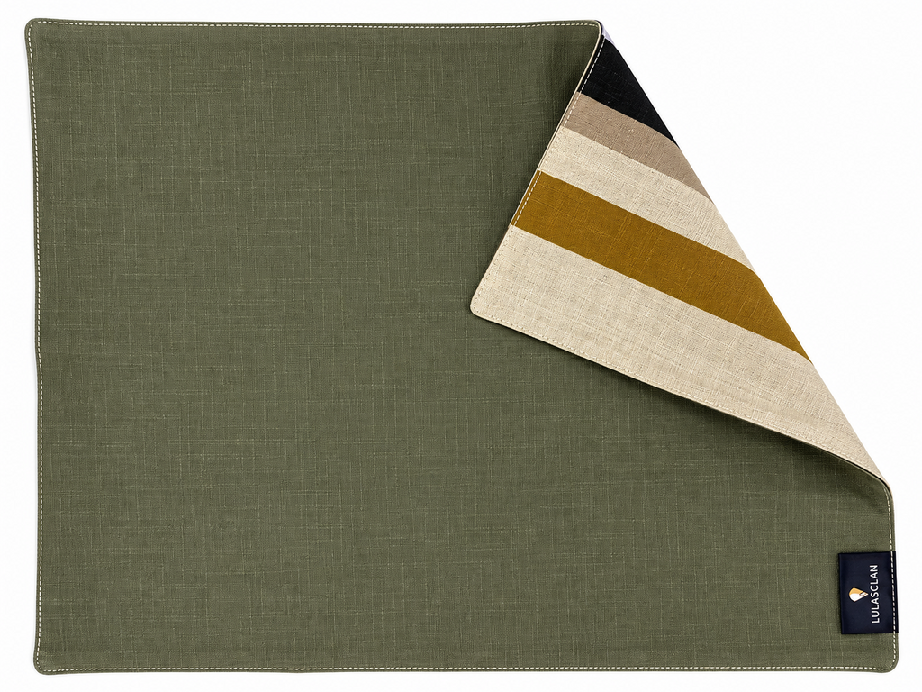 The Pastures placement by Lulasclan is shown with an olive green cotton ramie background fabric. A folded corner revealing a striped reverse side in beige, brown, and mustard yellow. A small black Lulasclan brand tag is visible in the bottom right corner.
